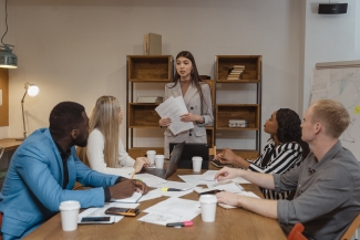 Standing woman speaks to four coworkers seated at a table